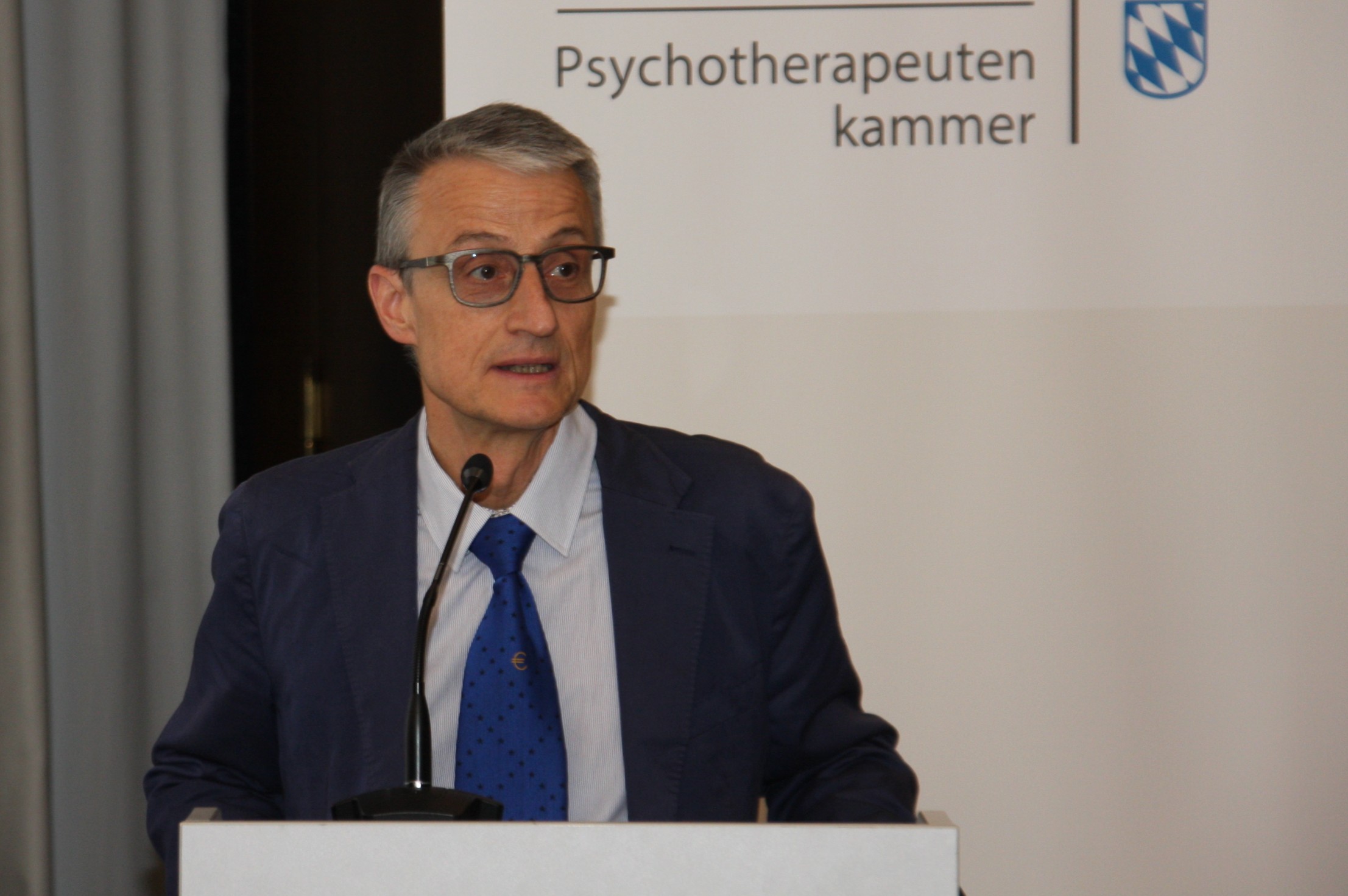 Ein Mann in einem dunkelblauen Anzug und Brille steht am Redepult. Im Hintergund ist ein Banner der PTK Bayern zu sehen.